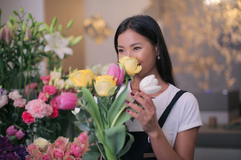 A young woman with long dark hair, smiling softly, holds a bouquet of fresh flowers close to her face in a flower shop. The bouquet features yellow tulips and pale pink roses, with green stems and lea