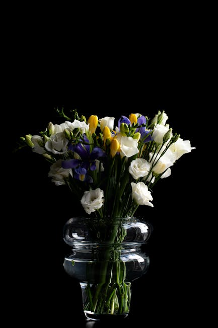 A floral bouquet arranged in a clear glass vase featuring a mix of fresh flowers, including white lisianthus, purple irises, and yellow tulips, with visible green stems and leaves. The blooms are arra