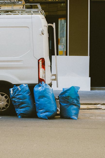 The image shows the rear side of a white delivery van parked on a street in front of a building with large windows and a glass door. Four large blue plastic bags, likely containing bulky floral waste,