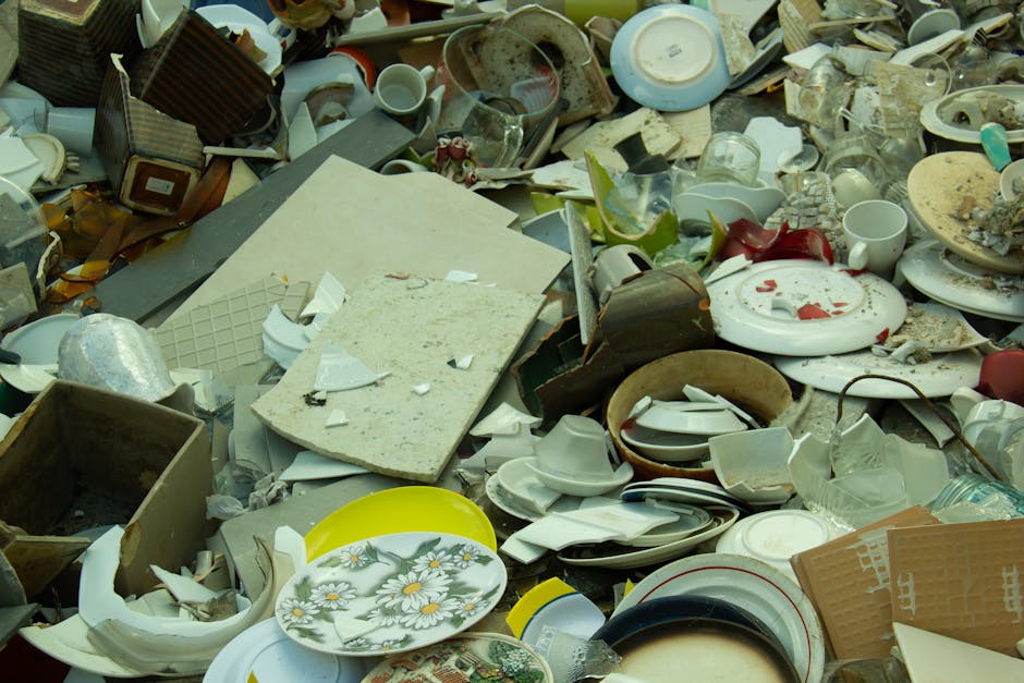 A large pile of bulky ceramic, glass, and plastic household waste, including broken plates with floral patterns, bowls, cups, and fragments of dishes, mixed with cardboard boxes, white foam, and vario