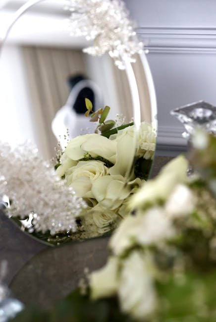 A close-up view of a floral arrangement featuring creamy white roses and delicate white calla lilies, accented with small green foliage, set against a reflective mirror. The bouquet is arranged in a s