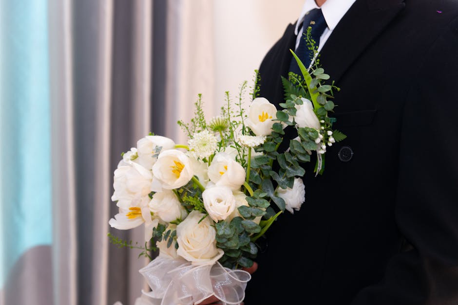 A close-up of a floral arrangement held by a person dressed in formal black attire, featuring a bouquet of white flowers including roses, calla lilies, and small blossoms, accented with green foliage 
