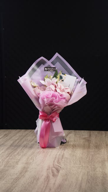 A bouquet arrangement of fresh flowers featuring soft pink roses, white lilies, and small sprigs of greenery, wrapped in light pink tissue paper and transparent cellophane, with a pink satin ribbon ti