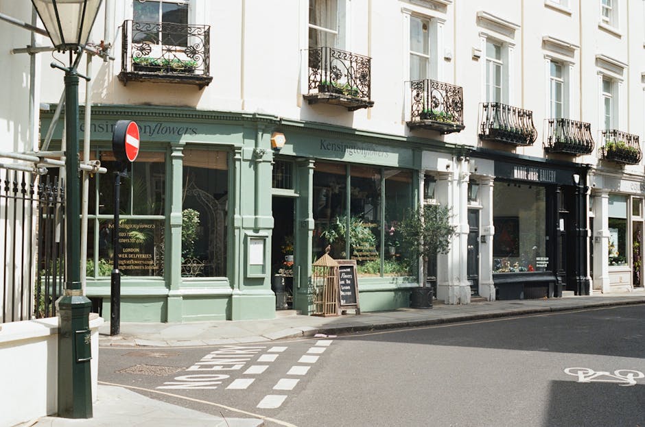 A storefront of a flower shop with a light green facade, large glass windows displaying various floral arrangements inside, and a small chalkboard sign outside. The shop has black decorative wrought-i