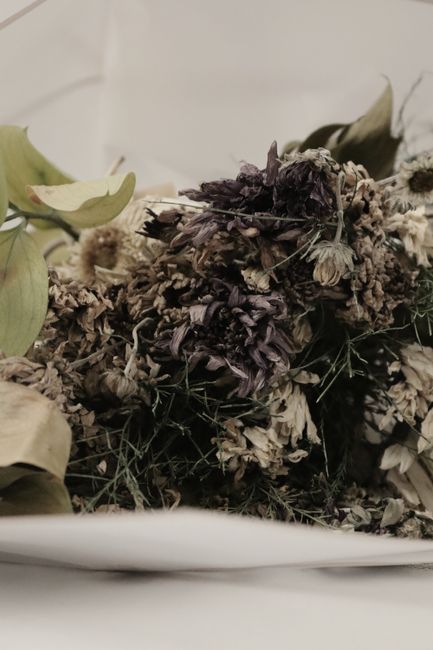 A close-up view of a collection of dried flowers arranged on a white surface, featuring faded purple, cream, and beige petals with hints of green foliage. The arrangement appears to be a floral gift o